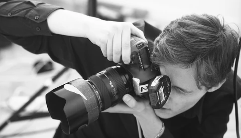 Photographer holding a DSLR camera with telephoto lens up to their face, shooting in black and white.