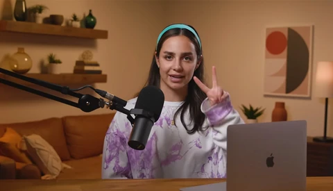 Woman speaking into a microphone at a desk with a laptop, making a peace sign gesture.