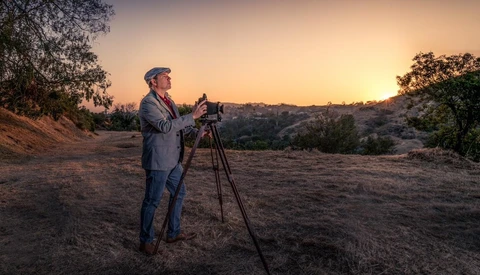 Surveyor in business casual attire operating a tripod-mounted surveying instrument during golden hour on a hillside.