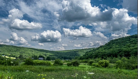 Rolling green hills with scattered farmhouses beneath a dramatic sky of white cumulus clouds.
