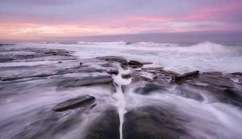 Waves crash over dark rocky outcrops during twilight with a pink and purple sky.