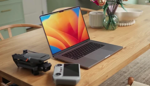 Laptop displaying colorful abstract wallpaper on wooden desk with drone and controller nearby.