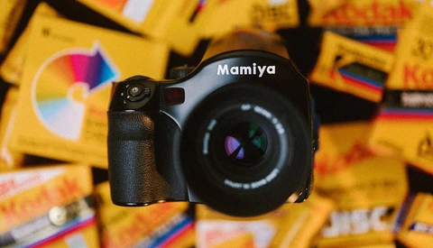 Mamiya medium format camera with colorful film boxes blurred in background.