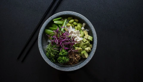 Overhead view of a gray bowl filled with sliced cucumber, edamame, purple cabbage, ginger, and seaweed on black surface.