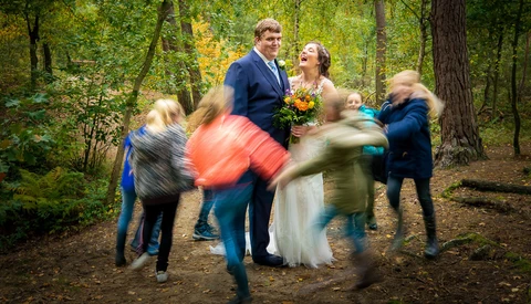 Bride and groom surrounded by blurred children running in a forest clearing during a wedding celebration.