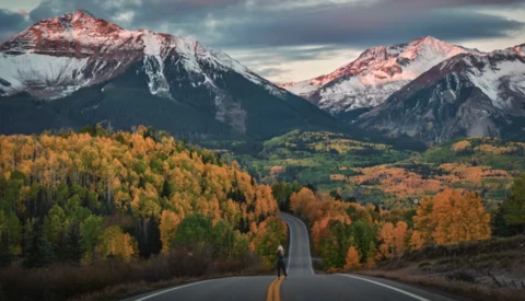 Straight road stretching toward snow-capped mountains framed by autumn forest in golden and orange tones.