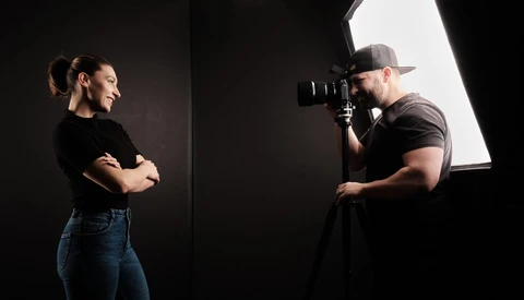 Photographer with camera on tripod shooting portrait of woman in studio with key light.