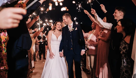 Bride and groom walk through a tunnel of sparklers held by wedding guests at night.