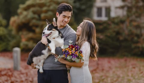 Young couple holding a husky dog and fall flowers outdoors in a brick-paved setting.