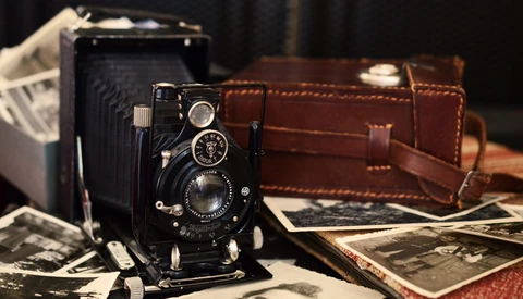 Vintage folding camera displayed with leather case, scattered black and white photographs, and another camera in soft studio lighting.