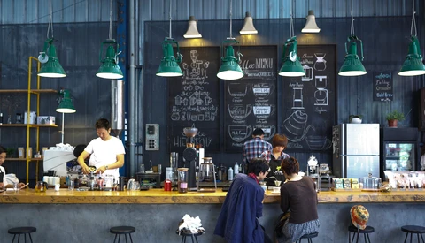 Modern café interior with customers at counter, green pendant lights, and chalkboard menu wall.