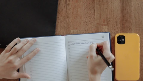 Person's hands writing in an open notebook with a pen, with a yellow smartphone visible on a wooden desk.
