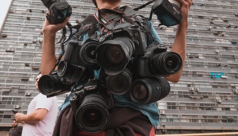 Photographer holding multiple Canon cameras and lenses against urban building backdrop.