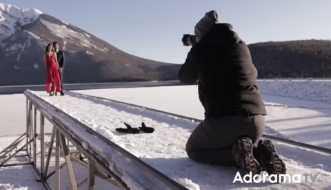 Photographer shooting a portrait of a woman in a red dress standing on a snowy dock, with snow-covered mountains and a frozen lake in the background.