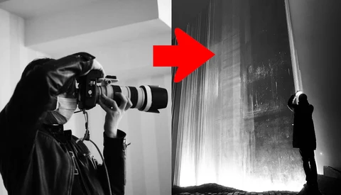 Photographer holding a camera with a telephoto lens while being photographed by another person in a minimalist interior space.