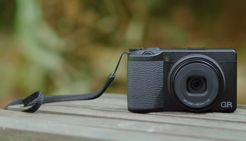 Ricoh GR compact camera with black wrist strap on wooden surface with blurred green foliage background.