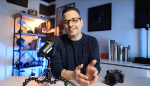 Content creator gesturing while speaking at a desk with a microphone, camera, and lighting equipment visible.