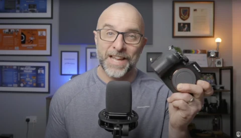 Man holding a camera lens toward the viewer while speaking into a microphone in a studio setting.