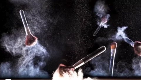Multiple makeup brushes suspended in mid-air with powder explosions against a dark background.