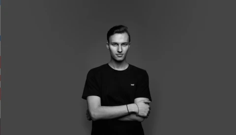 Young man in black t-shirt with arms crossed, standing against neutral gray background.