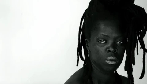 Black and white portrait of a person with braided hair looking directly at camera against plain white background.