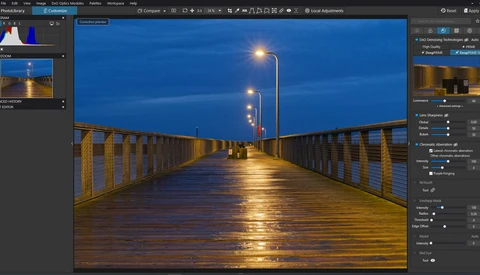 Long wooden pier at dusk with street lamps reflecting on wet surface under blue twilight sky.