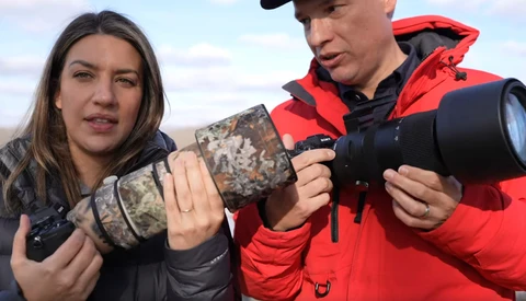 Two photographers holding telephoto lenses with camouflage and red jackets in an open landscape.