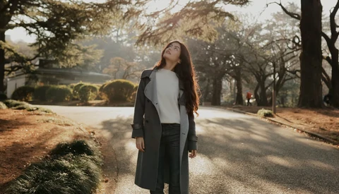 Woman in black coat looking upward while standing on a tree-lined path during golden hour.