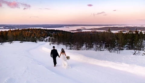 Bride and groom walking through snow-covered landscape at dusk with forested hills and frozen lake beyond.