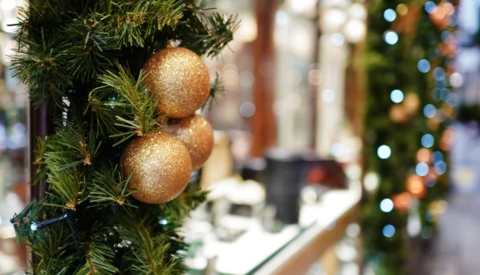 Two gold glitter ornaments hanging from evergreen branch with blurred holiday lights in background.
