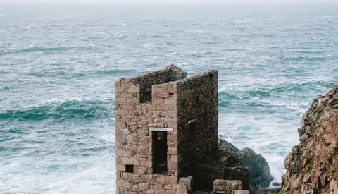Weathered stone tower ruins on a rocky coastal cliff overlooking turbulent ocean waves.