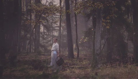 Woman in white shirt standing among tall trees in misty forest during autumn.