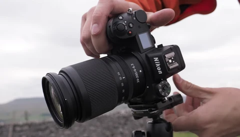 Hands holding a Nikon mirrorless camera with attached zoom lens against a blurred landscape background.