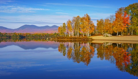 Autumn forest reflected in still lake water with mountains in the background.