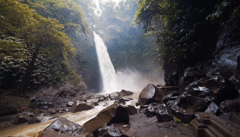 Waterfall cascading down a moss-covered cliff surrounded by dense jungle vegetation and scattered boulders.