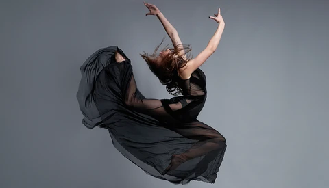 Dancer in mid-leap wearing black flowing fabric against neutral gray background.