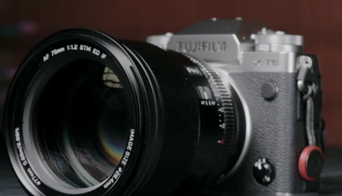 Fujifilm X-T5 camera with 75mm prime lens attached, shown in three-quarter view against a dark red background.