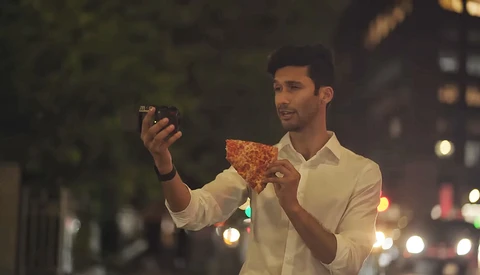 Man in white shirt holding pizza slice while taking selfie on city street at dusk.
