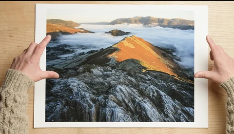 Printed landscape photograph held up against wooden background, showing golden-lit rocky peak above sea-level clouds with islands visible beyond.