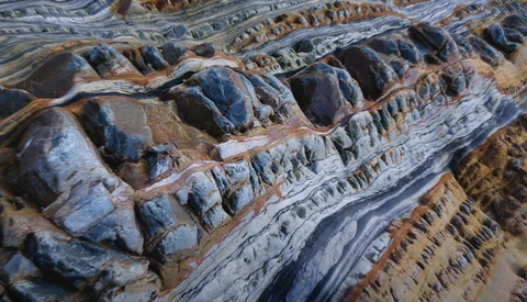 Aerial view of deeply eroded mountain terrain with sharp ridges and layered geological formations in warm earth tones.