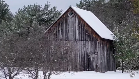 Weathered wooden barn with peaked roof surrounded by bare trees and snow.
