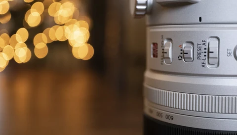Close-up of a camera lens showing focus ring, aperture, and autofocus controls with warm bokeh lights in background.