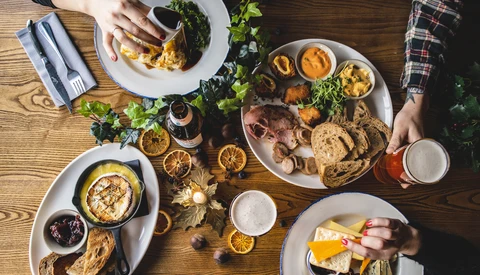 Overhead view of a shared meal with multiple white plates of food, fresh herbs, and beverages on a wooden table.