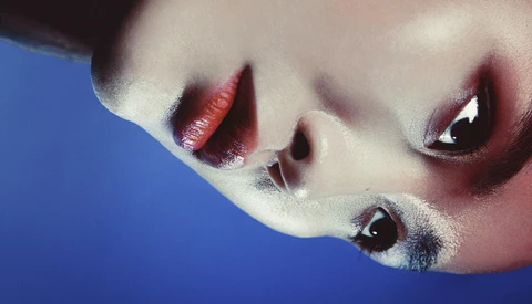 Close-up portrait of a face against blue background with bold red and burgundy lip makeup and dramatic eye shadow.