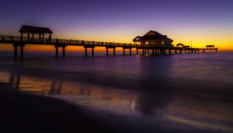 Silhouetted pier with buildings at sunset, reflected in wet sand and shallow water.