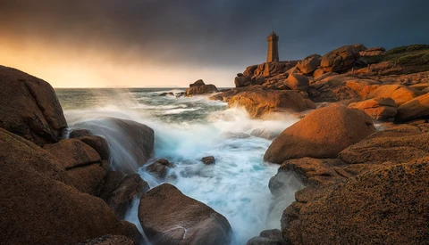 Lighthouse on rocky coastline at golden hour with waves crashing against granite boulders.