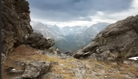 Alpine valley landscape framed by rocky peaks with dramatic cloudy sky and distant mountains.