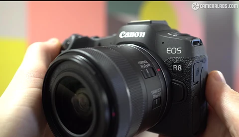 Canon EOS R8 mirrorless camera held in hands with attached lens against colorful background.
