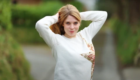 Young woman in white sweater posing outdoors with hands behind head, looking at camera.