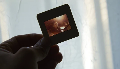 Hand holding a film slide transparency backlit against a bright sky, showing a warm-toned image within the frame.